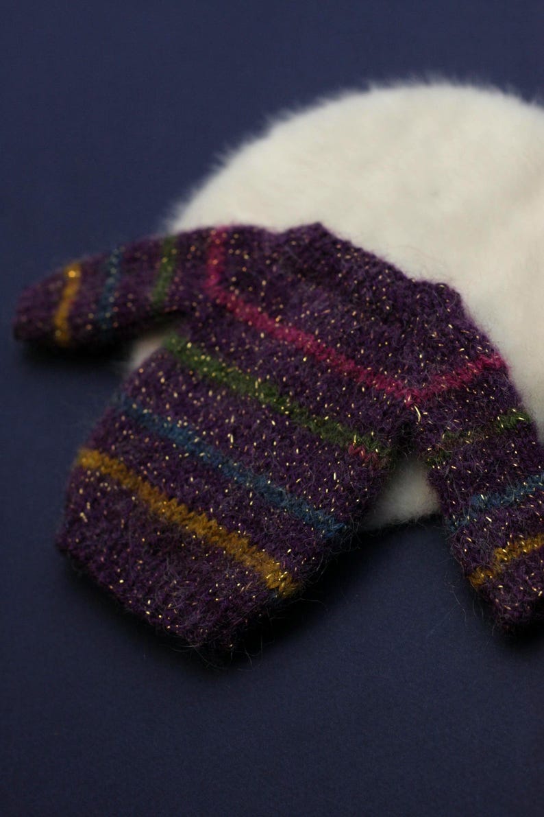 May include: A long-sleeved jumper crafted from fuzzy purple yarn with horizontal stripes in shades of green, blue, pink, and gold. The knit fabric features a subtle metallic gold thread woven throughout, creating a shimmering effect across the garment.