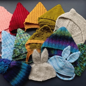 May include: A collection of handmade knit hats in various colors and styles. The hats include pointed elf-style hats in pink, red, yellow, and green, along with a gray bunny ear hat and a blue hat with a pom-pom. The hats are arranged on a dark surface.