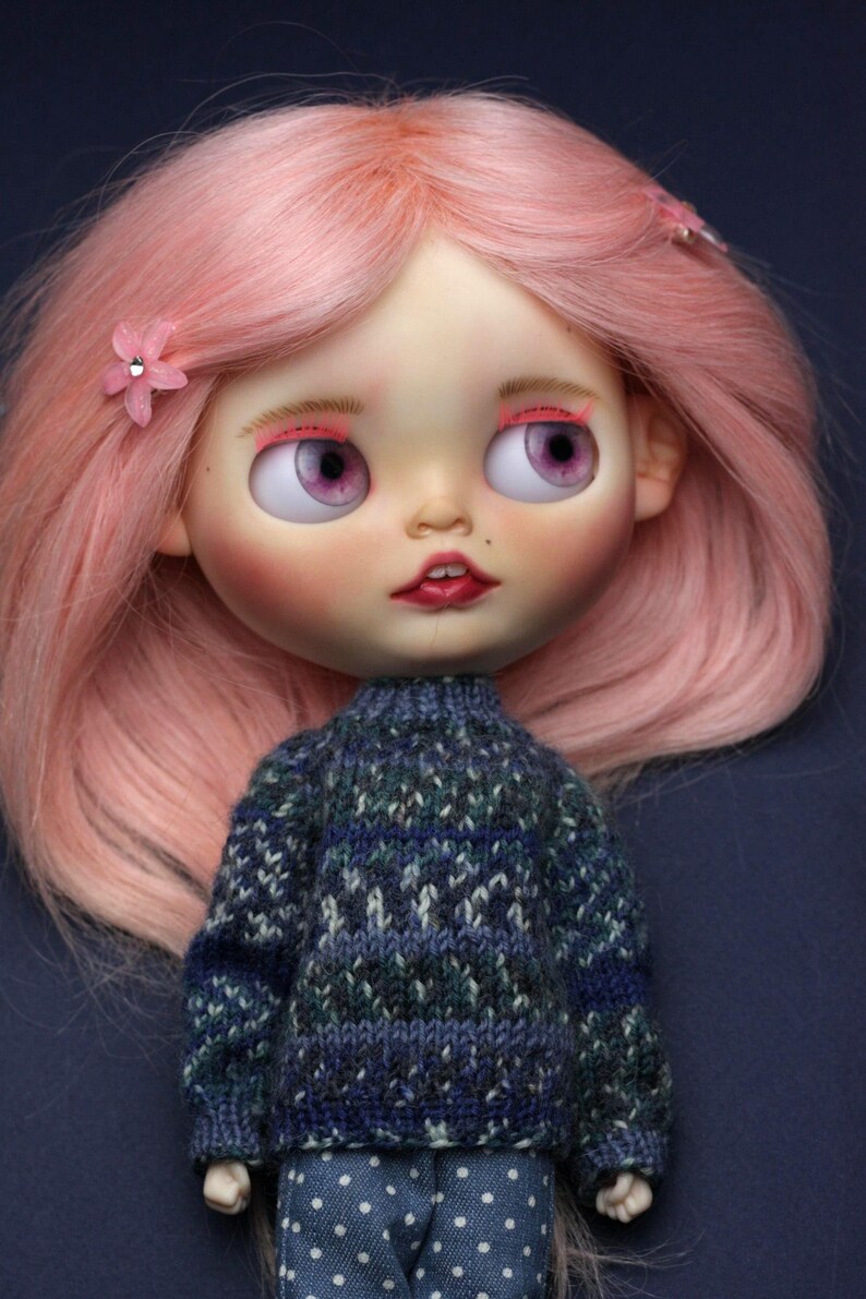 May include: A doll with long pink hair styled with small pink flower clips, featuring large purple eyes, pink eyeshadow, and a small beauty mark. The doll wears a dark blue and green patterned knit sweater over blue trousers with white polka dots.
