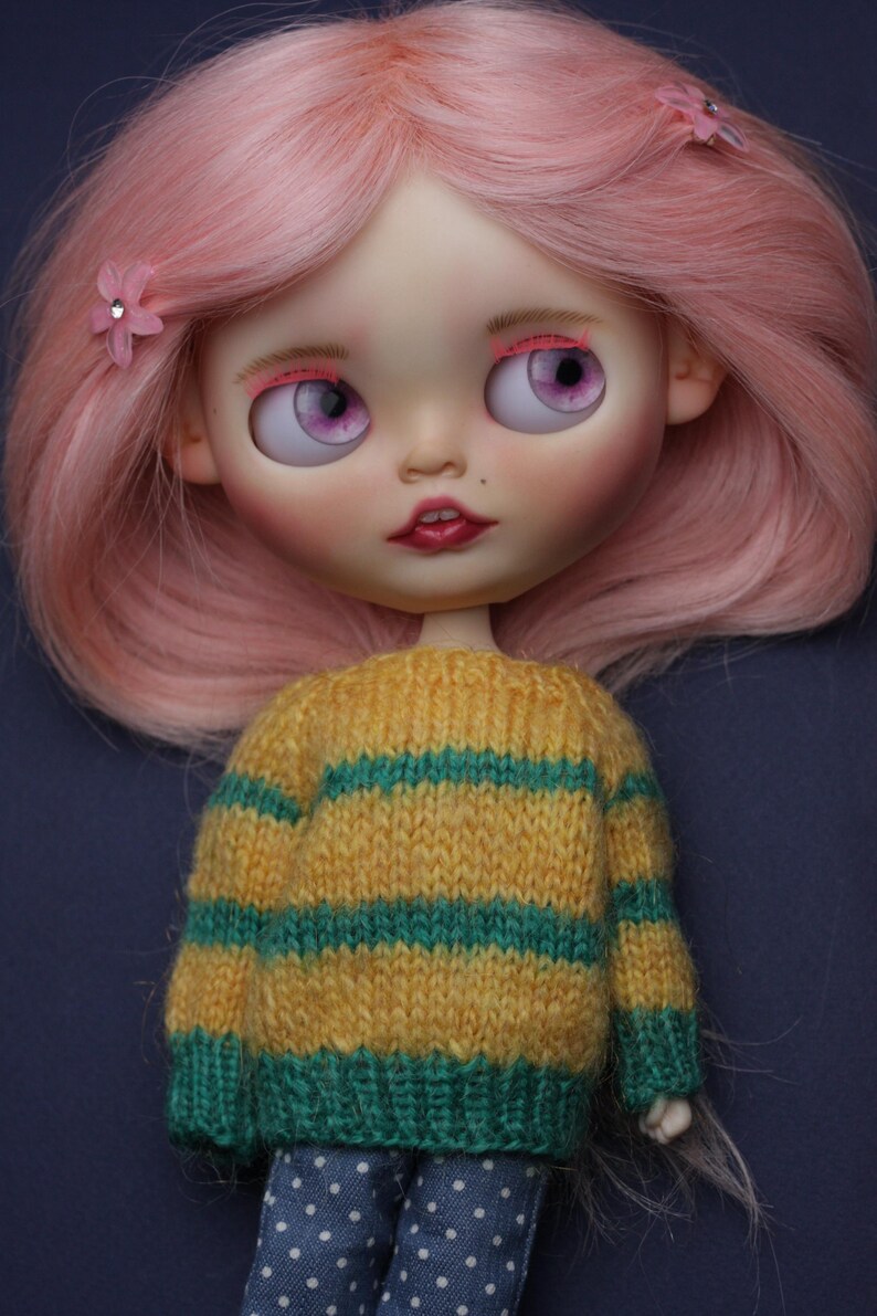 May include: A doll with shoulder-length pink hair, two small pink flower hair clips, and large purple eyes. The doll wears a yellow and green horizontal striped knitted jumper and blue trousers with a white polka dot pattern on a dark background.