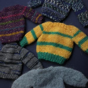 May include: A collection of five miniature knitted jumpers in various colours and patterns. The set includes a purple striped jumper with gold flecks, a blue and white patterned jumper, a yellow and green striped jumper, a grey striped jumper, and a solid light grey fuzzy jumper.