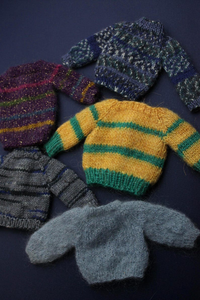 May include: A collection of five miniature knitted jumpers in various colours and patterns. The set includes a purple striped jumper with gold flecks, a blue and white patterned jumper, a yellow and green striped jumper, a grey striped jumper, and a solid light grey fuzzy jumper.