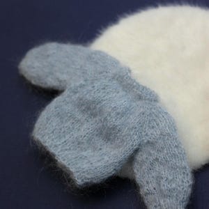 May include: A small, light blue knitted jumper with a fuzzy, textured surface, featuring long sleeves and a round neckline, positioned partially over a larger, round, white, fuzzy, and soft-textured knitted item.