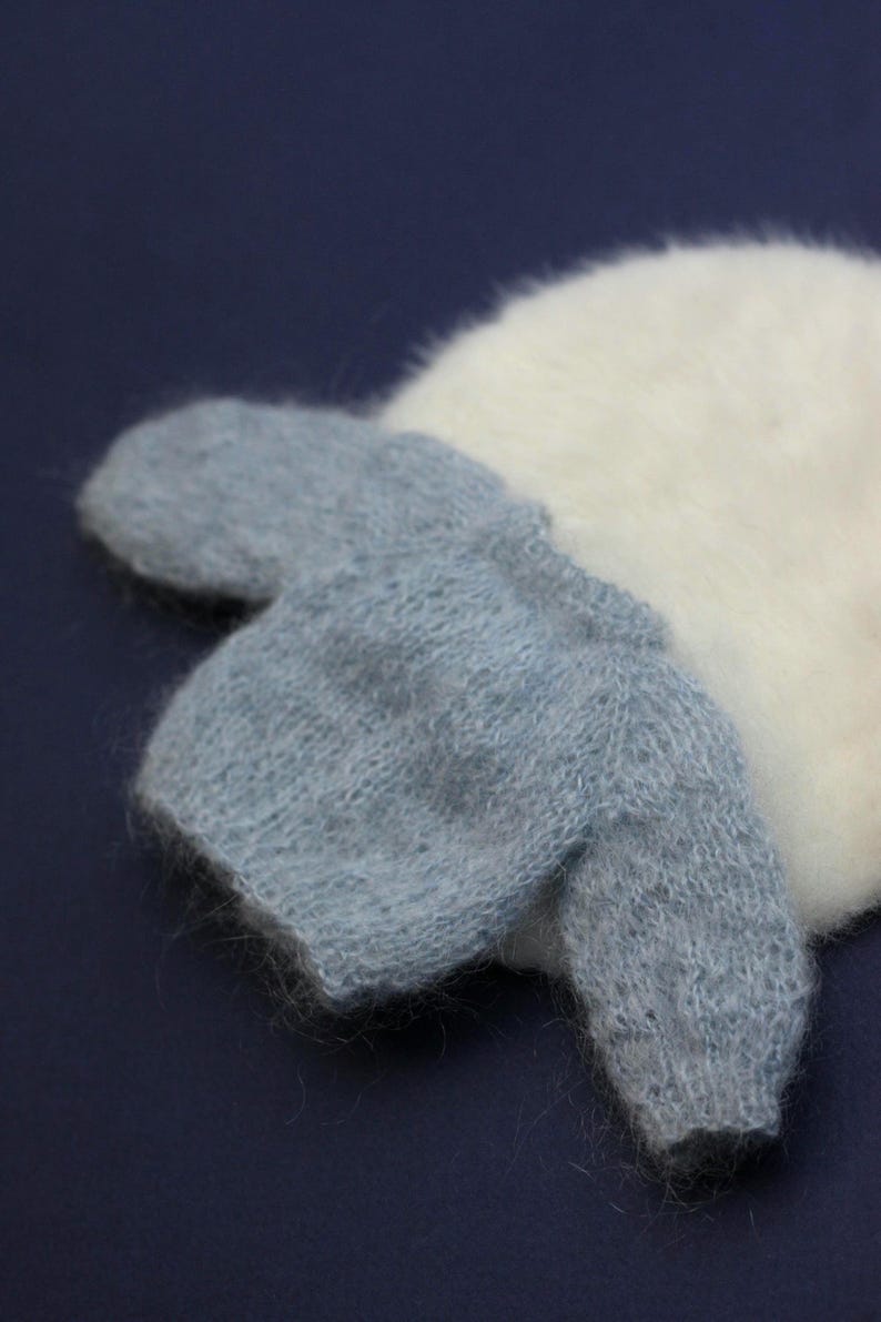May include: A small, light blue knitted jumper with a fuzzy, textured surface, featuring long sleeves and a round neckline, positioned partially over a larger, round, white, fuzzy, and soft-textured knitted item.