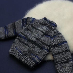 May include: A small, long-sleeved knit jumper featuring a horizontal striped pattern in shades of grey, black, and blue. The garment has a ribbed crew neckline, ribbed cuffs, and a ribbed hem, constructed with a visible yarn texture throughout.