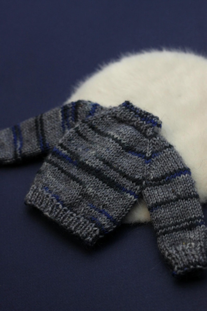 May include: A small, long-sleeved knit jumper featuring a horizontal striped pattern in shades of grey, black, and blue. The garment has a ribbed crew neckline, ribbed cuffs, and a ribbed hem, constructed with a visible yarn texture throughout.