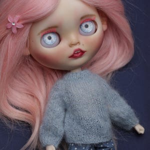 May include: A doll with long pink hair, pale skin, and large light blue eyes. The doll wears a grey knitted long-sleeve jumper and blue trousers with white polka dots. A small pink flower accessory with a clear rhinestone centre is in the hair.