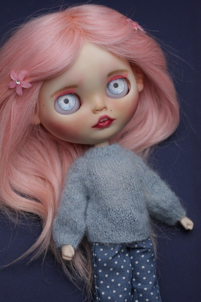 May include: A doll with long pink hair, pale skin, and large light blue eyes. The doll wears a grey knitted long-sleeve jumper and blue trousers with white polka dots. A small pink flower accessory with a clear rhinestone centre is in the hair.
