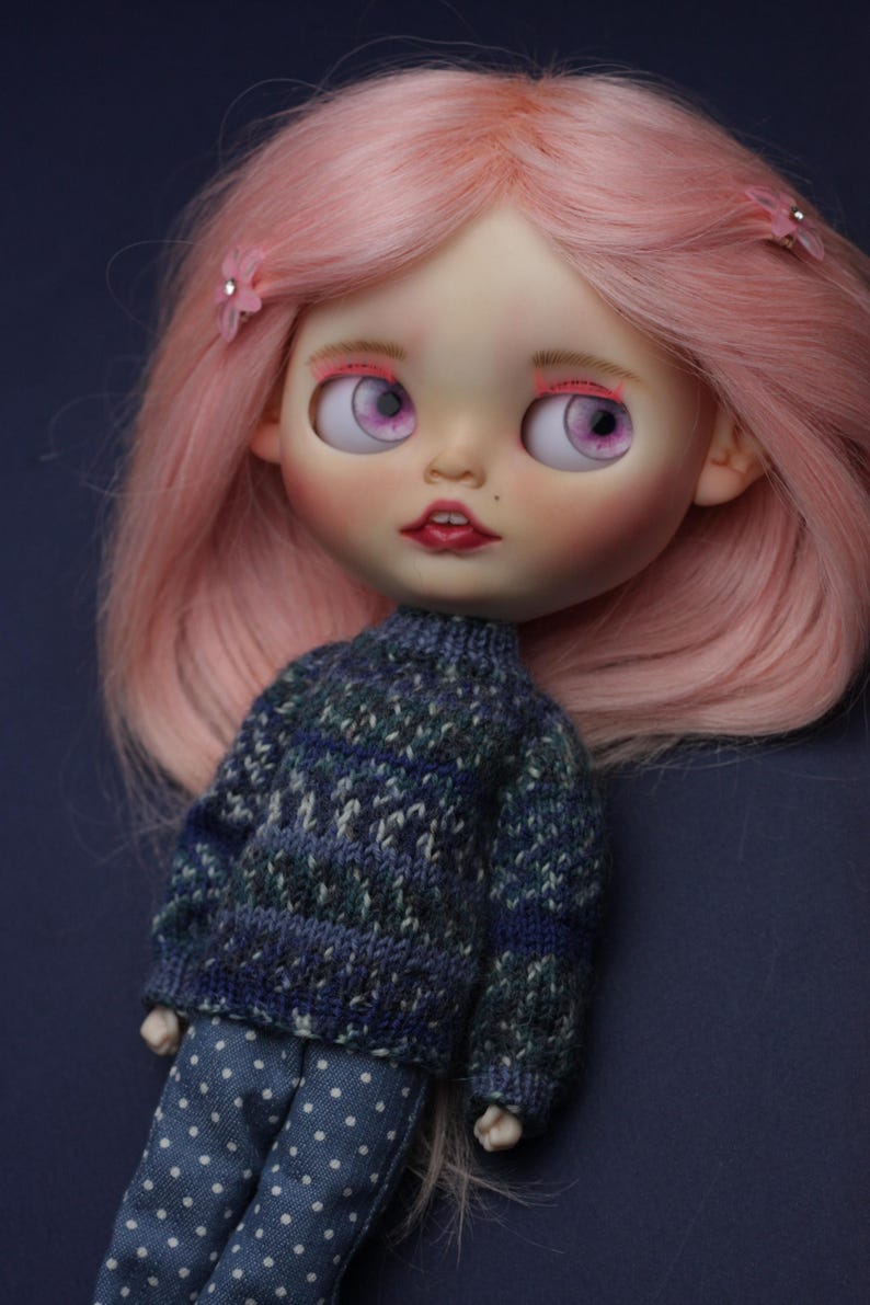 May include: A doll with long pink hair styled with two small pink flower clips. The doll has large light purple eyes, pink eyeshadow, and red lips. It wears a blue and green patterned knit jumper and blue trousers with a white polka dot pattern.