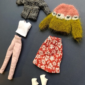 May include: A collection of doll-sized clothing items, including a patterned sweater, a white tank top, pink striped pants, a red floral skirt, a pink and green sweater, and white sneakers. The outfits are arranged on a dark blue surface.