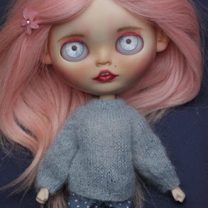 May include: A doll with long pink hair styled with two small pink flower clips. The doll has large pale blue eyes, pink eyebrows, and red lips. It wears a fuzzy light grey long-sleeved sweater and dark blue trousers featuring a white polka dot pattern.