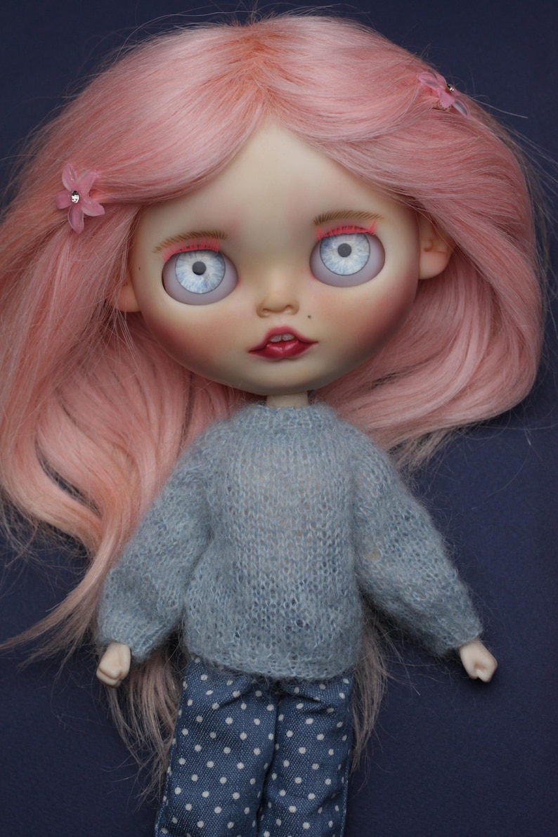 May include: A doll with long pink hair styled with two small pink flower clips. The doll has large pale blue eyes, pink eyebrows, and red lips. It wears a fuzzy light grey long-sleeved sweater and dark blue trousers featuring a white polka dot pattern.