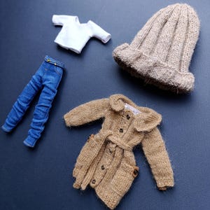Peut inclure: Ensemble de vêtements miniatures comprenant un t-shirt blanc, un jean bleu, un manteau en tricot beige avec une ceinture et un bonnet assorti. Le manteau a des boutons et des poches. Les articles sont disposés sur une surface bleu foncé.