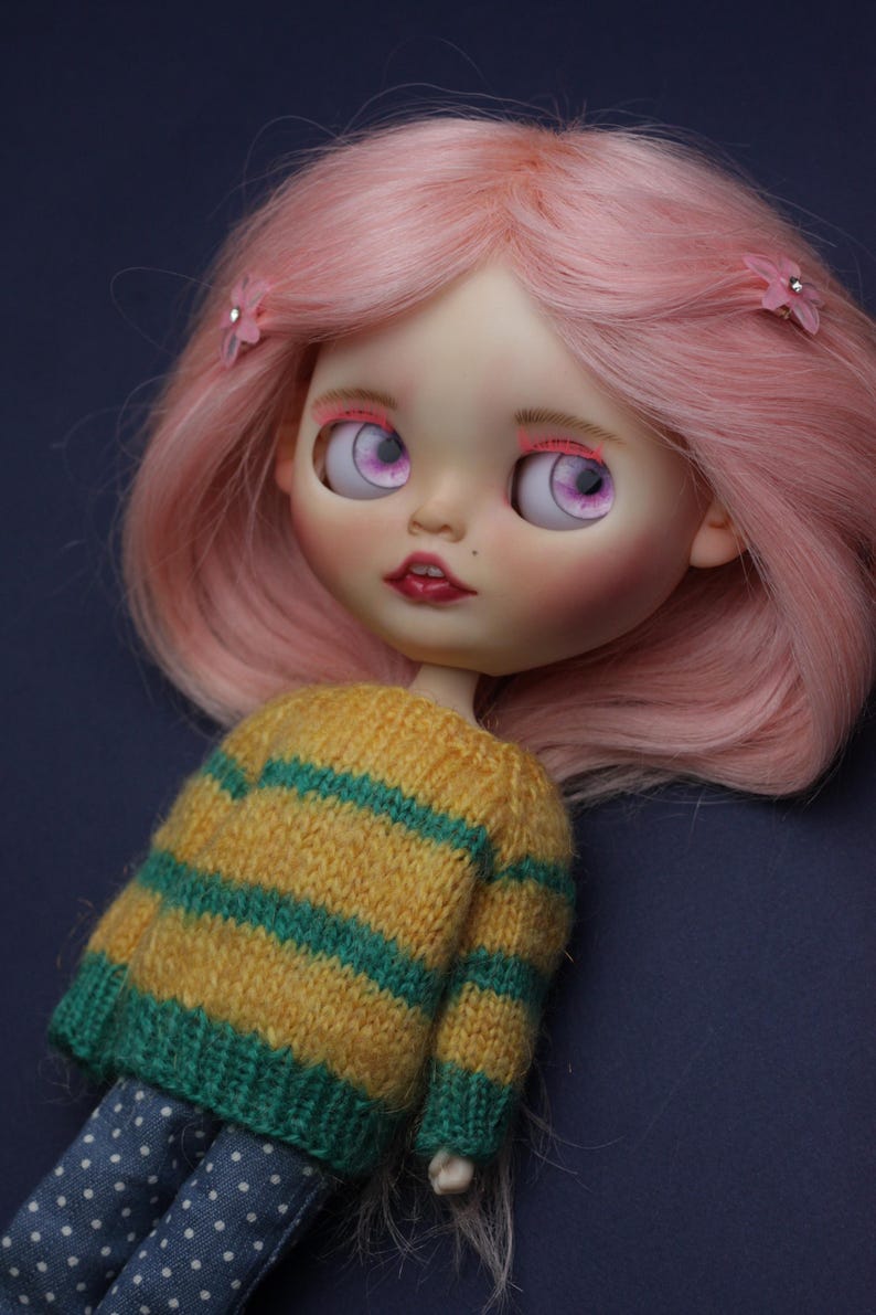 May include: A doll with shoulder-length pink hair styled with two small pink flower clips. The doll has large light purple eyes, pink eyeshadow, and dark red lips. It wears a yellow and green striped knit sweater and blue polka-dot trousers.