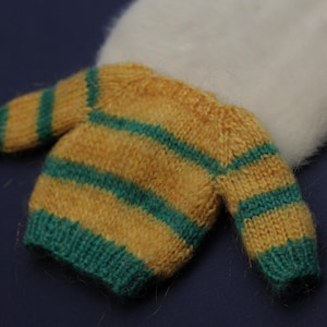 May include: A small, fuzzy, long-sleeved jumper featuring horizontal stripes in mustard yellow and teal green. The garment has a ribbed hem and cuffs, and is positioned in front of a soft, white, circular, plush, fuzzy material.