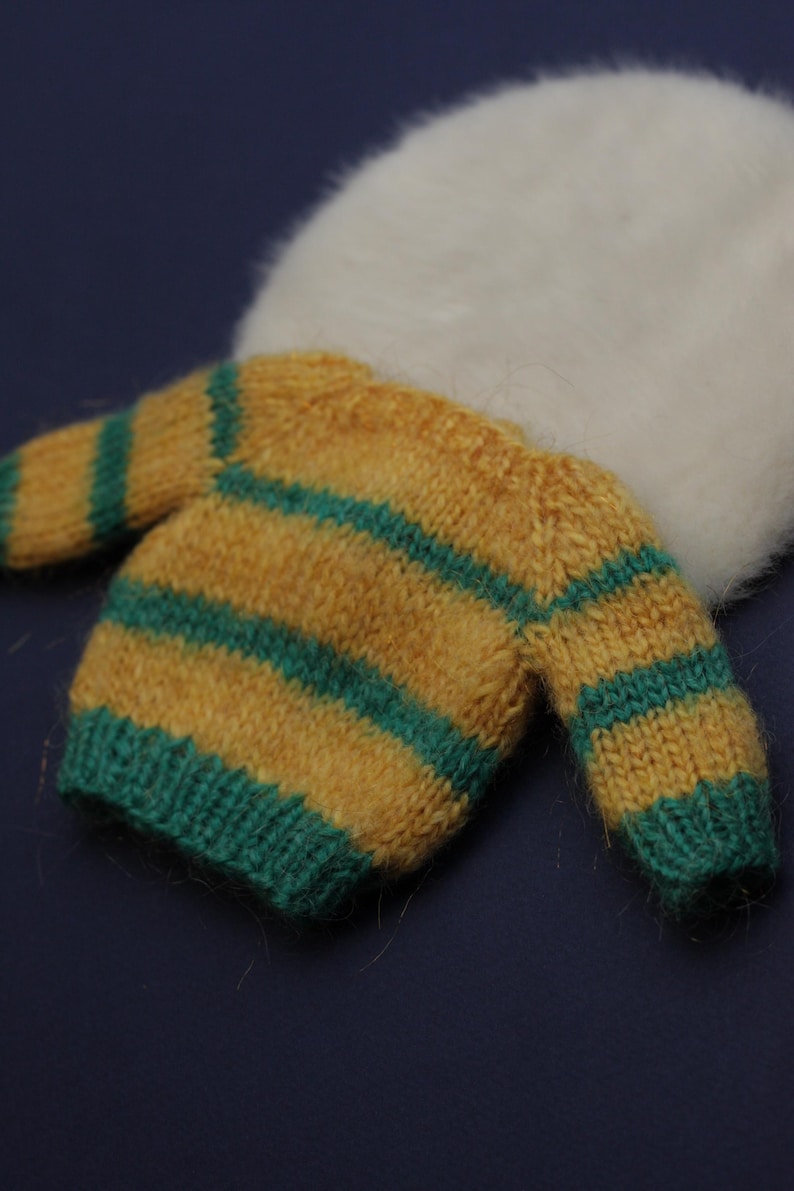 May include: A small, fuzzy, long-sleeved jumper featuring horizontal stripes in mustard yellow and teal green. The garment has a ribbed hem and cuffs, and is positioned in front of a soft, white, circular, plush, fuzzy material.