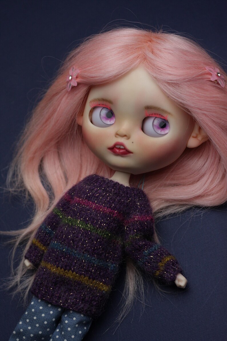 May include: A doll with long pastel pink hair styled with two small pink flower clips. The doll features large light purple eyes, pink eyelashes, and dark red lips. It wears a long-sleeved purple knit sweater with horizontal stripes and blue polka-dot trousers.