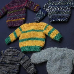 May include: Five miniature knitted jumpers in various colours and patterns. One is purple with horizontal stripes, one is blue with a textured pattern, one is yellow with green stripes, one is grey with dark stripes, and one is solid light grey.