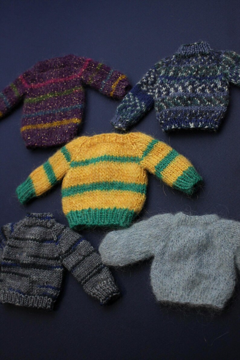 May include: Five miniature knitted jumpers in various colours and patterns. One is purple with horizontal stripes, one is blue with a textured pattern, one is yellow with green stripes, one is grey with dark stripes, and one is solid light grey.