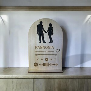 Könnte beinhalten: Eine Holzplakette mit einer Silhouette eines Paares, das sich an den Händen hält. Die Plakette trägt den Text "Pannonia" und "Desh x Young Fly x Azahriah" sowie ein Spotify-Logo und eine Musikplayer-Leiste mit dem Songtitel und der Zeit.
