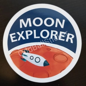May include: A white circle sticker with a blue background and the text "MOON EXPLORER" in white. A red planet with a white rocket ship is in the bottom half of the sticker.