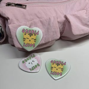 May include: Three heart-shaped badges with the words "stupid idiot" and a cartoon tiger or cloud. The badges are white with a green and pink gradient background. One badge is attached to a pink bag.