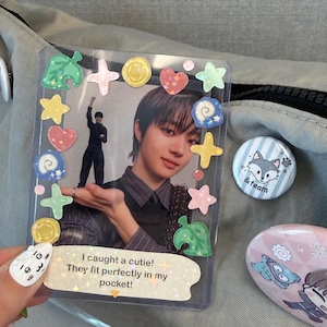 May include: A clear plastic sleeve holding a photo of a person, decorated with colorful stickers. The photo has the text "I caught a cutie! They fit perfectly in my pocket!" A round button with a wolf design and a pink oval-shaped object are also visible.
