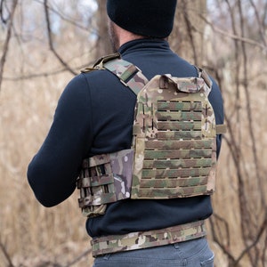 May include: A person wearing a tactical vest with a camouflage pattern. The vest has a large panel of MOLLE webbing on the back and a smaller panel on the front. The vest is secured with a belt.
