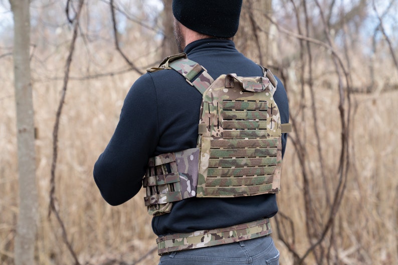 May include: A person wearing a tactical vest with a camouflage pattern. The vest has a large panel of MOLLE webbing on the back and a smaller panel on the front. The vest is secured with a belt.