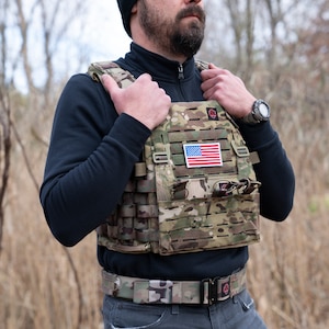 May include: A camouflage tactical vest with a velcro patch featuring the American flag. The vest has multiple pouches and straps for carrying gear.