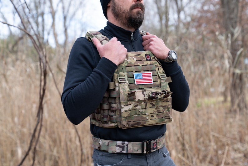 May include: A camouflage tactical vest with a velcro patch featuring the American flag. The vest has multiple pouches and straps for carrying gear.