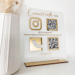 Könnte beinhalten: Ein weißes Acrylschild mit goldfarbenen Buchstaben und Akzenten. Auf dem Schild steht "Connect with us" und es enthält ein Instagram-Logo, ein Logo mit einem "C" und zwei QR-Codes. Das Schild steht auf einem goldenen Sockel.