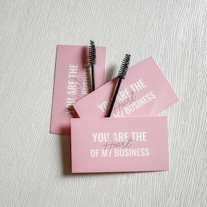 Può includere: Tre carte rosa con il testo "You are the Heart of my Business" impilate l'una sull'altra. Due applicatori di ciglia neri sono sopra le carte.