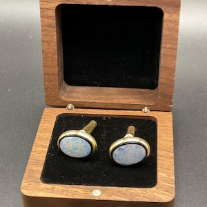 May include: A pair of oval cufflinks with a gold-colored border and a light blue and white opal-like center, presented in a wooden box with a black velvet interior. The box is open, revealing the cufflinks.