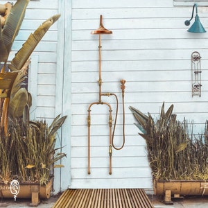 May include: A copper shower system featuring a round shower head, a vertical pipe, and a handheld shower attachment with a flexible hose. The unit includes brass valves and connectors, with two parallel pipes extending downward for water supply connections.