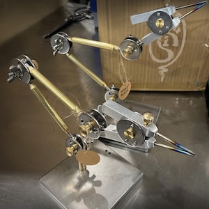 May include: Several metal fly tying vises with brass and silver components are displayed. Each vise has a clamp, adjustable arms, and a small tag that says "Hand Made". The vises are on a metal surface.