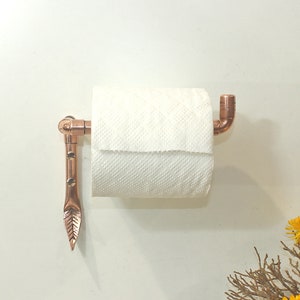 Könnte beinhalten: Ein Toilettenpapierhalter aus Kupfer mit einem blattförmigen Arm und einer weißen Rolle Toilettenpapier.
