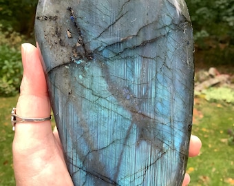May include: A large, polished, blue labradorite gemstone with iridescent flashes of color. The stone is shaped like an oval and has a smooth, polished surface.