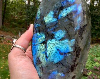 May include: A large, freeform, polished labradorite crystal with a blue and green iridescent sheen. The crystal is held in a hand, showcasing its size and unique shape.