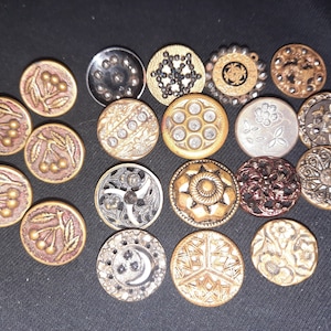 Op de afbeelding: Een verzameling vintage metalen knopen in verschillende ontwerpen en kleuren, waaronder goud, zilver en rood. De knopen hebben bloemmotieven, geometrische en abstracte patronen.