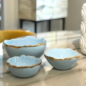 May include: Three light blue ceramic bowls with gold rims. The bowls are nested inside each other, with the largest bowl at the bottom and the smallest bowl at the top.