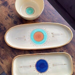 Könnte beinhalten: Drei Keramik-Servierplatten mit goldenen Rändern. Die Platten sind weiß mit einem kreisförmigen Design in der Mitte. Die mittleren Designs sind türkis und orange, türkis und orange und blau.
