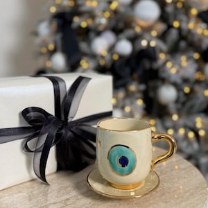 May include: A white ceramic coffee mug with a gold rim and a blue and green evil eye design. The mug is sitting on a matching saucer.