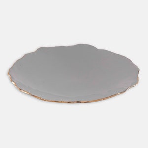 May include: A grey, irregularly shaped ceramic plate with a gold-coloured rim. The plate has a smooth surface and a slightly textured edge. Suitable for serving appetizers or small dishes. Measures approximately 25 cm in diameter.