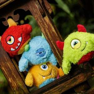 May include: Four plush toys shaped like monsters, each with a different color and expression. The toys are arranged in a stack, with the red monster on top, followed by the blue monster, then the yellow monster, and finally the green monster on the bottom.