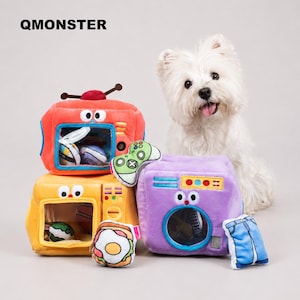 May include: A white dog sits behind three plush toys: an orange television, a yellow microwave, and a purple washing machine. The toys feature cartoon eyes, clear windows, and small plush accessories including a game controller, toast, and blue jeans. The text QMONSTER appears in the top left corner.