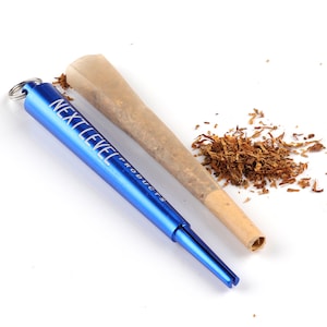 Puede incluir: Una herramienta de enrollado de metal azul con un llavero y el texto "Next Level Products" en el lateral. Un cono de papel marrón está al lado de la herramienta, y hay un montón de tabaco suelto en el fondo.