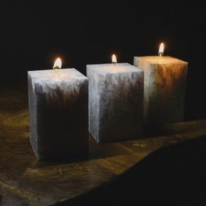 May include: Three grey square candles with white marbling are lit and placed on a dark wooden surface.
