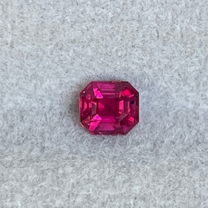 May include: A vibrant red gemstone with a rectangular emerald cut. The gemstone is set against a white background.