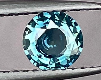 Può includere: Una pietra preziosa rotonda e sfaccettata di colore blu-verde. La pietra preziosa è tenuta da una pinzetta su uno sfondo grigio.
